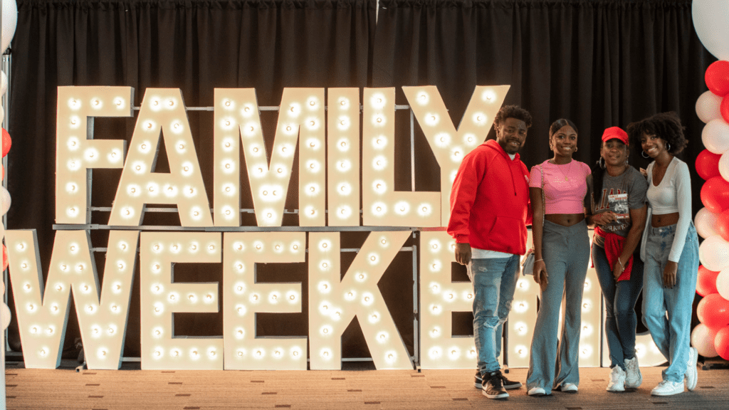 A family of four standing in from of a lighted Family Weekend sign