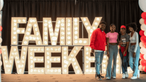 A family of four standing next to a glamour lighted "Family Weekend" sign 