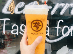 A person holding up an orange drink in front of a sign.