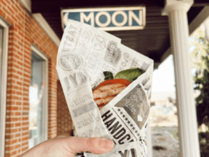 Store front image of Moon Co-op with a wrapped sandwich held in the center