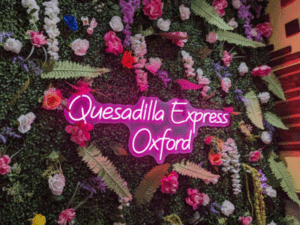Neon sign for "quesadilla express mexican restaurant oxford" surrounded by a decorative floral wall.