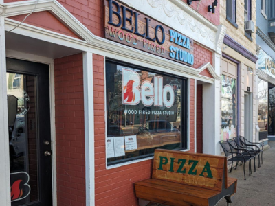 A storefront of Bello Wood Fired Pizza Oxford studio with a bench and sign out front.