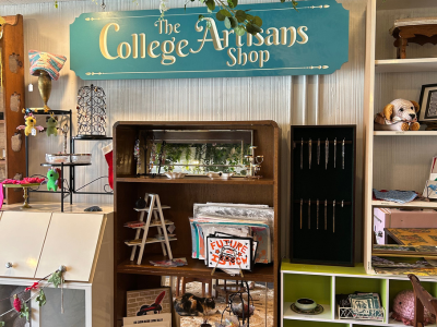 A wooden blue sign inside the shop says The College Artisans Shop with items for sale below