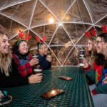 A group of friends wearing antler headbands sit inside a garden igloo on a picnic table with hot cocoa