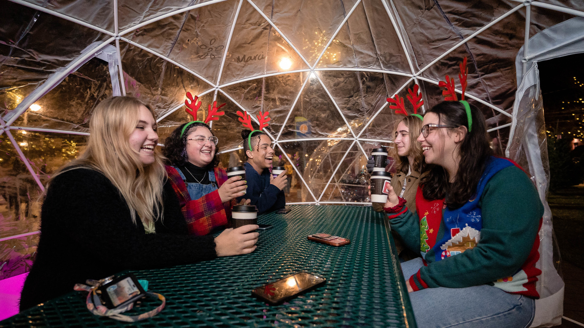 A group of friends wearing reindeer headbands inside a garden igloo on a picnic table with cups of hot cocoa
