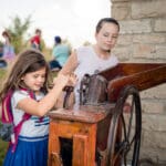 A young girl turns the hand crank on a pioneer tool as her friend watches.