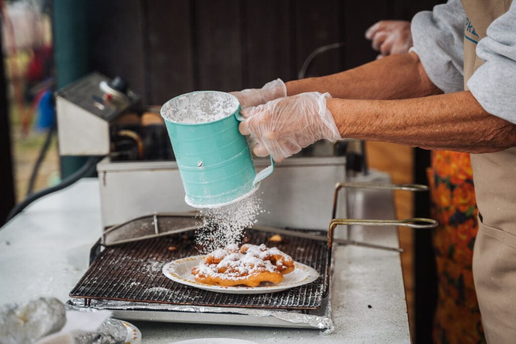 A funnel cake sits on a rack after being pulled from the fryer. Someone is topping it with powdered sugar.