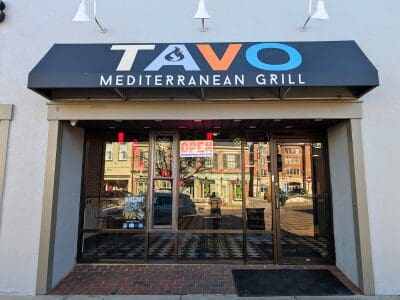 Light grey exterior bricks with black canopy shop sign with multi-colored letters spelling "TAVO" above glass windows and entrance door.