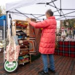 A woman stands at a local vendor's booth, holding up and examining a handmade rug.