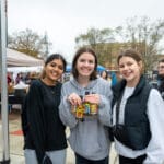 Three young women pose in the middle of Uptown Oxford. The middle woman is holding three bottles of hot sauce.