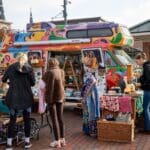 Event attendees stand in front of a colorfully decorated van, sorting through various garments and other items available for purchase.
