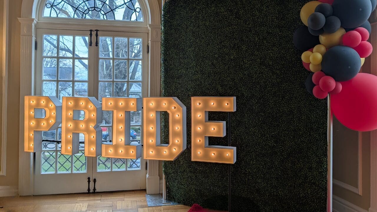 Golden marquee letters that spell "PRIDE" in front of a window and selfie station