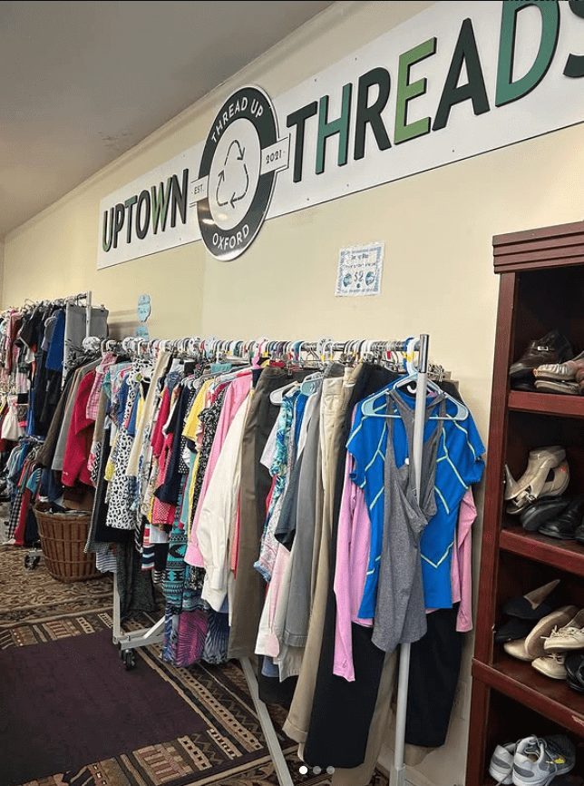 Photo of a wall sign "Uptown Threads" with a recycle icon in the middle placed above a rack of secondhand clothing