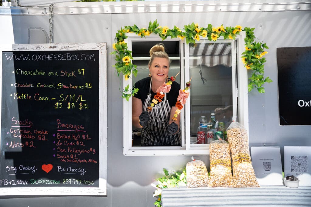 A woman leans out the window of a food truck, showcasing the kabobs she's selling.