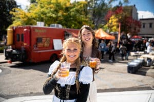 Two women dressed in traditional German garb pose for the camera with beers in hand.