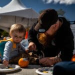 A father and his young child sit on the ground in front of a painted pumpkin.
