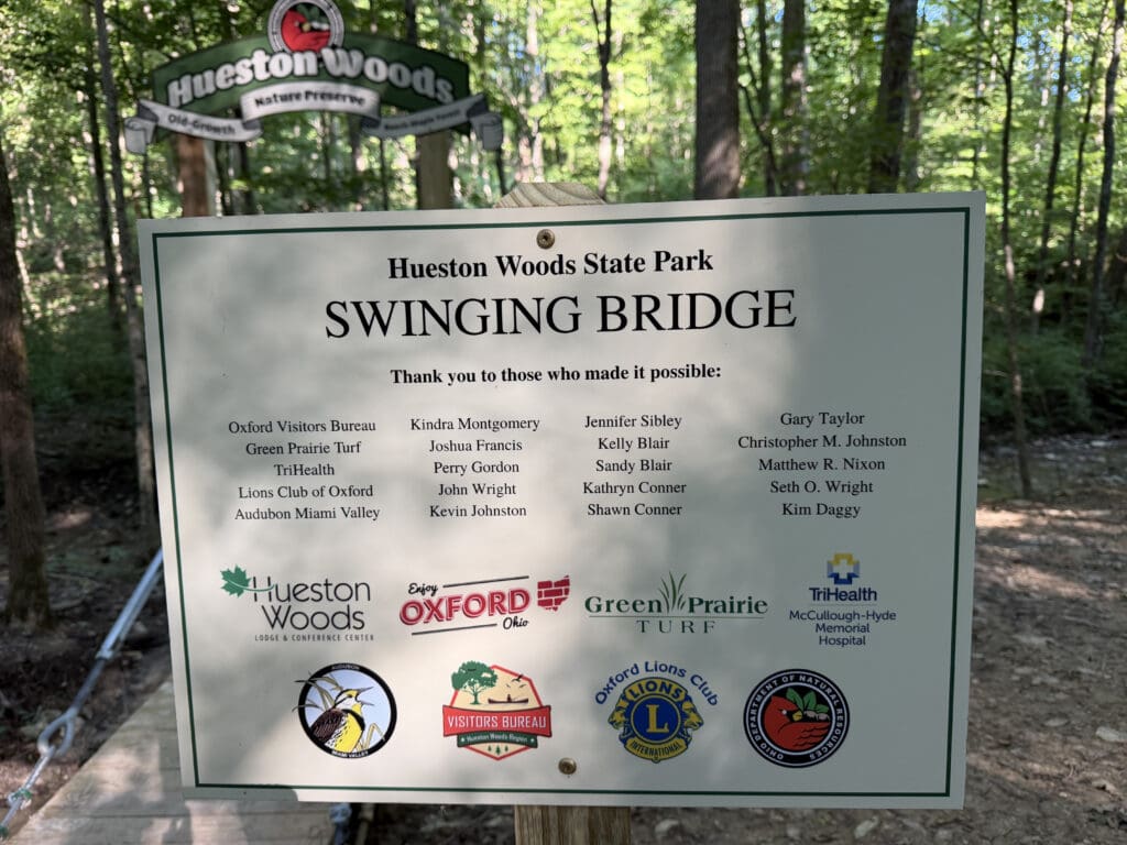 A sign with the following text: Hueston Woods State Park / Swinging Bridge / Thank you to those who made it possible: Oxford Visitors Bureau, Green Prairie Turf, TriHealth, Lions Club of Oxford, Audubon Miami Valley, Kindra Montgomery, Joshua Francis, Perry Gordon, John Wright, Kevin Johnston, Jennifer Sibley, Kelly Blair, Sandy Blair, Kathryn Conner, Shawn Conner, Gary Taylor, Christopher M. Johnston, Matthew R. Nixon, Seth O. Wright, Kim Daggy. After that text, are the following logos: Hueston Woods Lodge and Conference Center, Enjoy Oxford, Green Prairie Turf, TriHealth McCullough-Hyde Memorial Hospital, Audubon Miami Valley, Visitors Bureau Hueston Woods Region, Oxford Lions Club, Ohio Department of Natural Resources