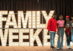 A family of four standing in from of a lighted Family Weekend sign