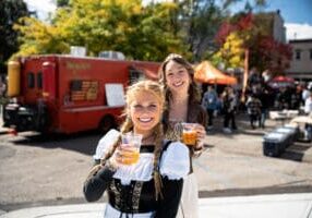 Two women dressed in traditional German garb pose for the camera with beers in hand.